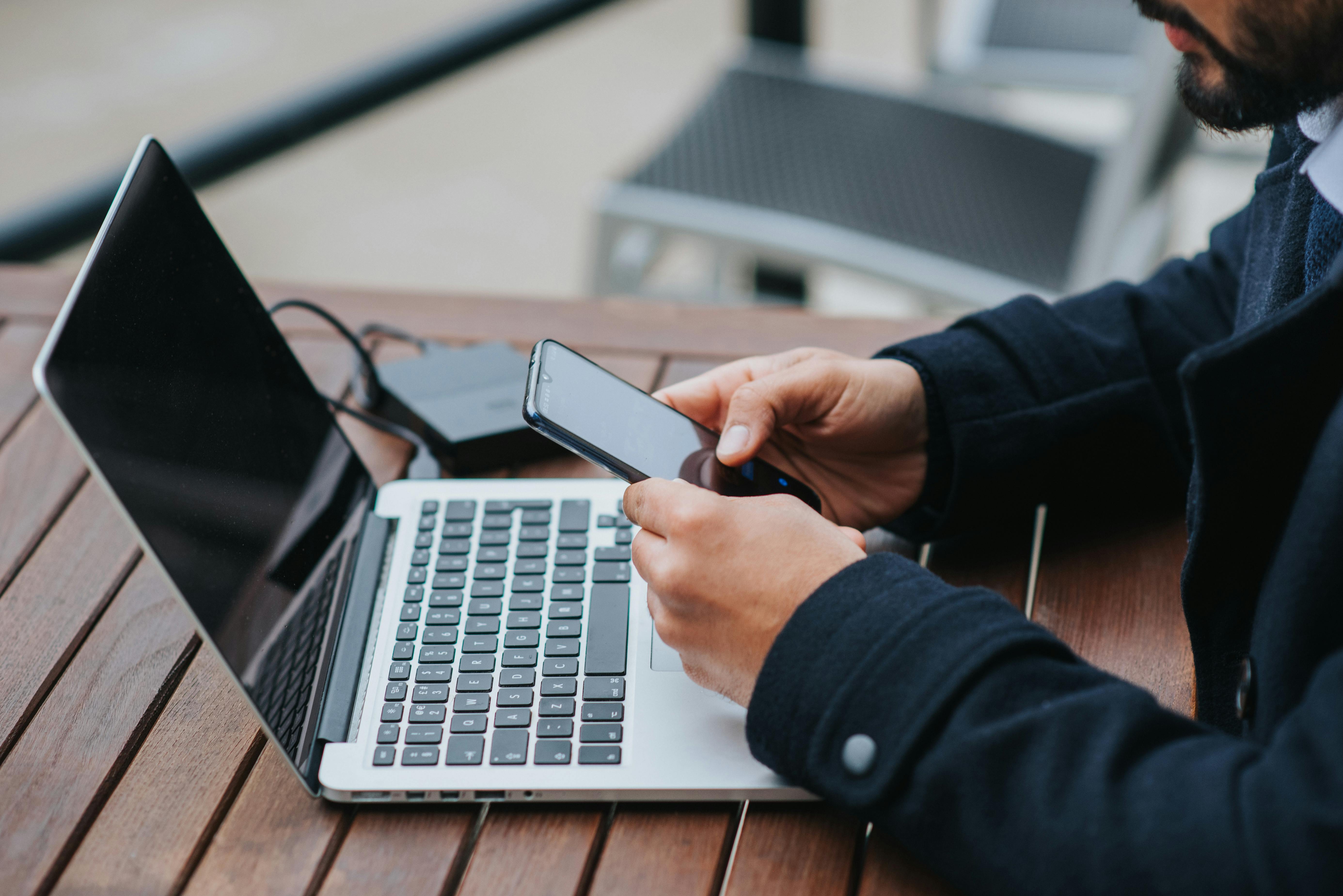 Homem jovem de sobretudo usando o celular, com um notebook sobre a mesa